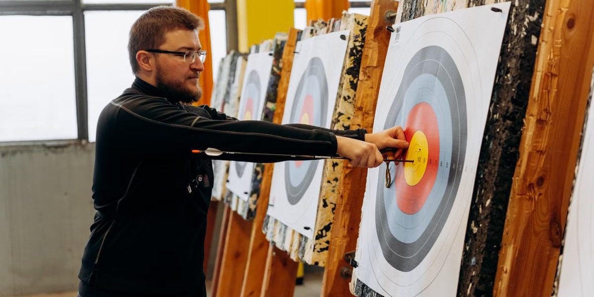 Man in glasses practicing archery indoors, pulling arrows from bullseye on target.