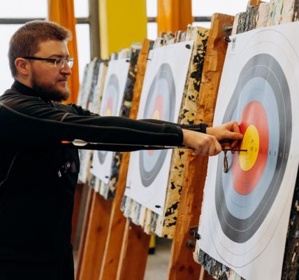Man in glasses practicing archery indoors, pulling arrows from bullseye on target.