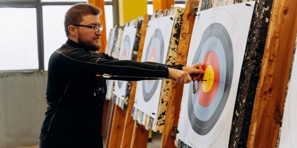 Man in glasses practicing archery indoors, pulling arrows from bullseye on target.